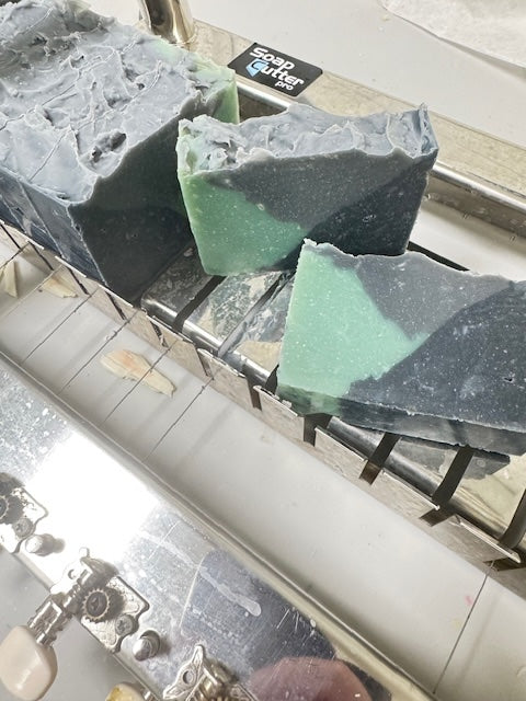 Two green and gray soap bars on a metal surface newly cut from a loaf. 