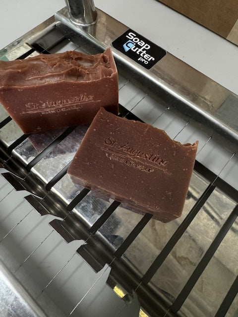 Two bars of soap on a metal soap cutter with 'St. Augustine Bath & Candle' branding.